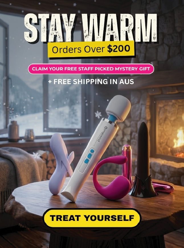 An advertisement banner for an adult novelty product, showing a white and blue wand massager, a pink curved insert, and a black flared tip resting on a wooden table in front of a cozy fireplace. Prominent text reads 'STAY WARM Orders Over $200' and 'TREAT YOURSELF'.