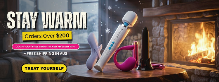 An advertisement banner for an adult novelty product, showing a white and blue wand massager, a pink curved insert, and a black flared tip resting on a wooden table in front of a cozy fireplace. Prominent text reads 'STAY WARM Orders Over $200' and 'TREAT YOURSELF'.