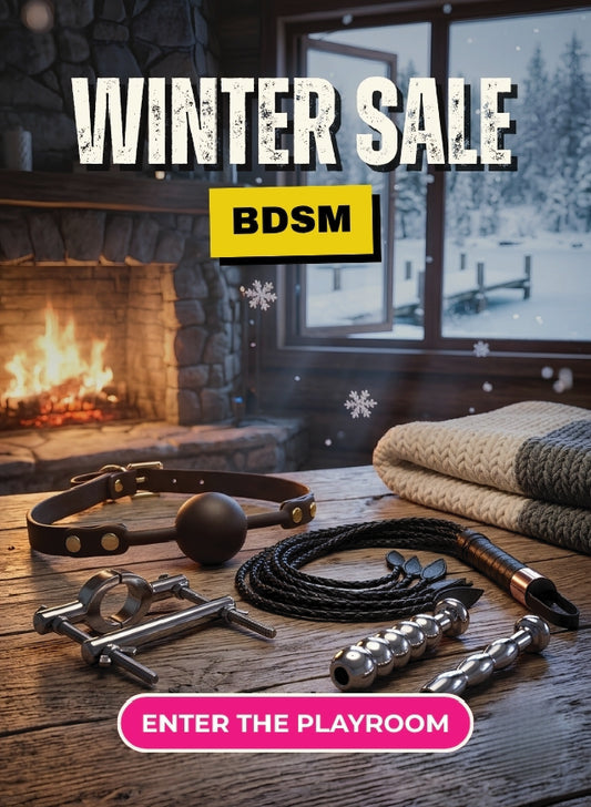 A promotional winter sale graphic showing BDSM play items arranged on a wooden table in front of a cozy fireplace and snowy window. Visible items include a leather collar with a dark spherical insert, metal bars and hardware, a coiled black rope, a textured strap or harness, and folded knit blankets.